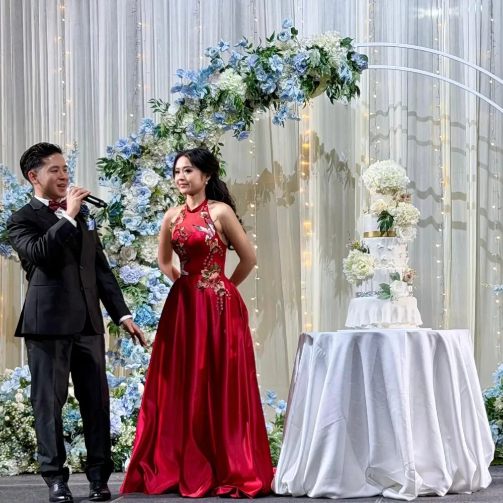 BRIDE WEARING A CUSTOM RED CHEONGSAM-NECKLINE TEA CEREMONY GOWN WITH ORIENTAL FLORAL EMBROIDERY, STANDING BESIDE A WEDDING CAKE